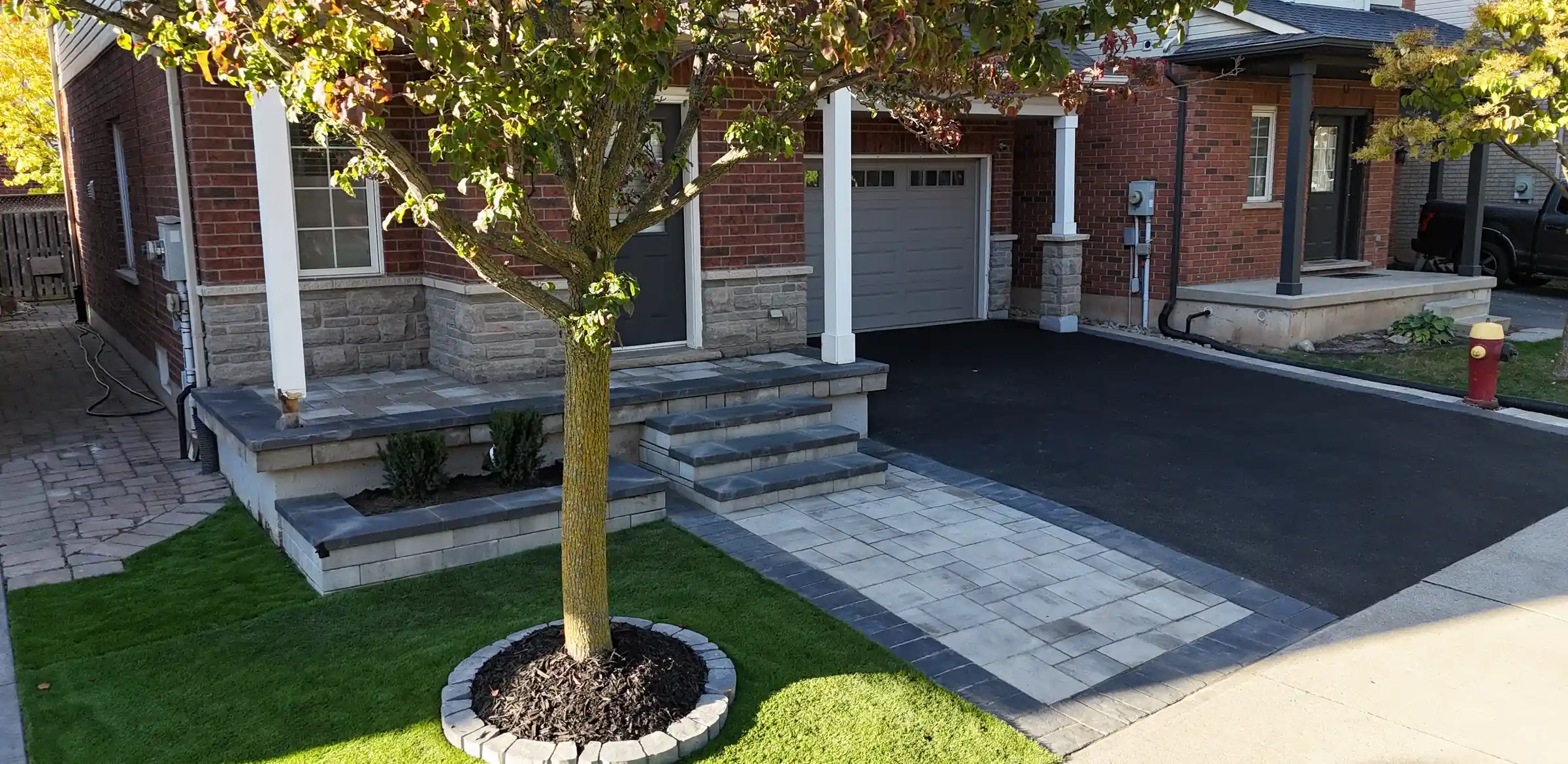 Interlock walkway and steps with a stone-ringed tree bed in front of a brick home