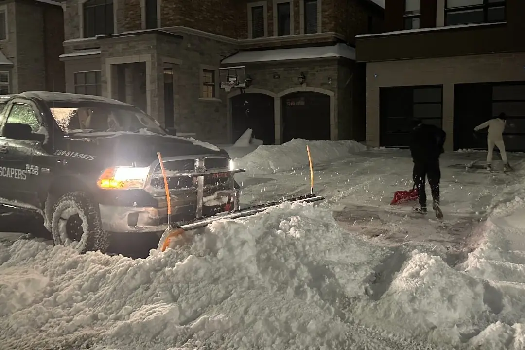 MJ plow truck clearing a driveway at night while crew shovel