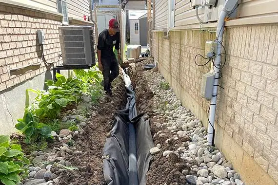 French drain being installed in a narrow side yard lined with river rock